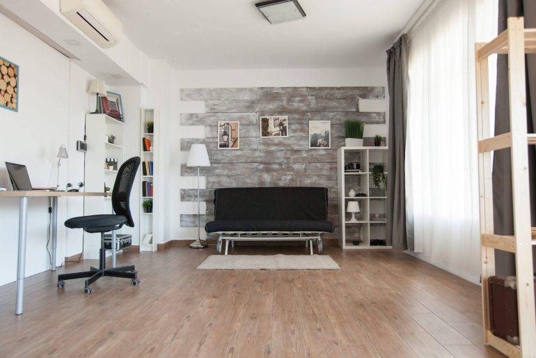 Bright modern living area and home office with a desk, laptop, white shelving, and a gray distressed-wood feature wall behind a black sofa.